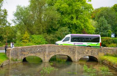 A Go Cotswolds minibus crossing a quaint bridge over a river in a leafy English village.