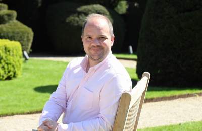 An image of Liam Bartlett, Chair of Shakespeare's England sitting in a chair outdoors in manicured gardens and smiling at the camera.