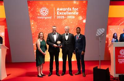 Representatives from Warwick Conferences pose on stage at the VisitEngland Awards for Excellence 2025–2026 ceremony, holding a tourism award in front of a brightly lit orange backdrop displaying the event branding. Four formally dressed attendees stand on a red carpet during the awards presentation.