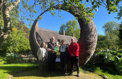 Team members from the Shakespeare Birthplace Trust pose with their Green Tourism accreditation award in the gardens of Anne Hathaway’s Cottage, standing beneath a large woven willow sculpture on a sunny spring day.