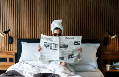 A woman sitting in bed wearing sunglasses, a white dressing gown with a white towel turban on her head reading the Telegraph newspaper.