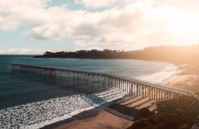 Long wooden pier stretches over blue ocean to a sandy beach at sunset, with warm sunlight, gentle waves and a California coast.
