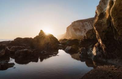 Sunset over a rugged California coastline with a calm tidal pool reflecting towering cliffs and a starburst of light between rocks.