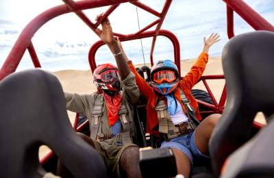 Two people with helmets and goggles in a dune buggy with hands up
