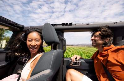 Two people smiling with open top car with wind in hair and fields in background