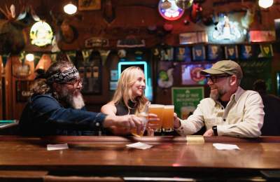 Three seated people cheersing will beer at a wooden bar with illuminated beer signs, flags and decor on the wall behind