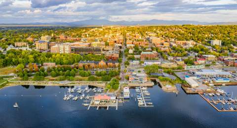 Aerial view of Burlington by a lakeshore with marinas, docks, and numerous buildings. Greenery surrounds the area, and mountains are visible in the background.