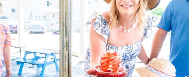 A woman in a large brim hat is handed a strawberry sundae out the window of an ice cream shop.