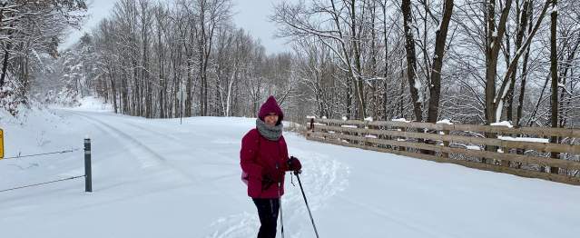 A woman is bundled up in winter gear and wearing corss country skis with poles in hand at a snow-covered trail head.