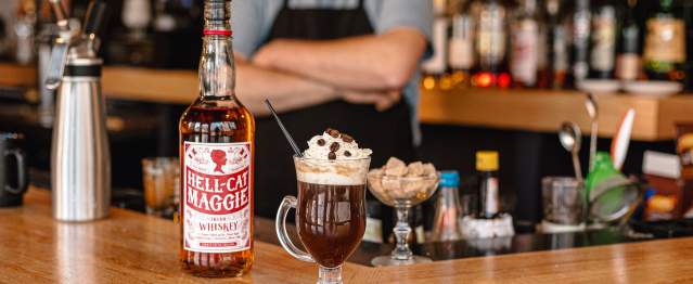A coffee drink with whipped cream and coffee beans sits on a bar next to a bottle of Hell Cat liquor.