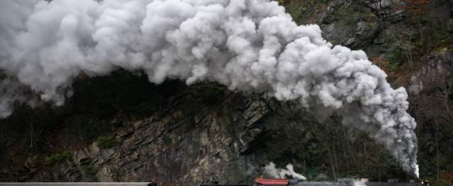 The Western Maryland Scenic Railroad steam engine billows smoke from the engines and pulls along a passenger car near a mountain wall.