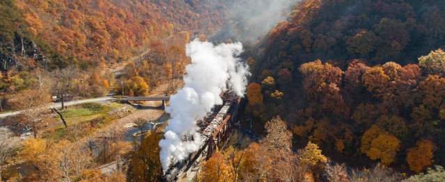 A steam train travels through a mountain valley surrounded by vibrant autumn foliage.