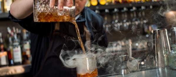 Bartender pouring a smoking Cedar cocktail at a bar with aromatic presentation