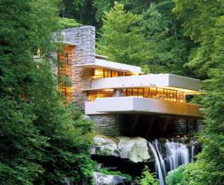 A modern home with a balcony and a waterfall coming out of the bottom. Green luscious trees are surrounding the architectural marvel.