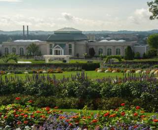 Exterior of a large conservatory sounded by grounds of lush green trees and bushes and flower beds with colorful florals