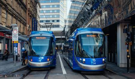 Two blue trams, parked side by side in a street, let passengers embark.