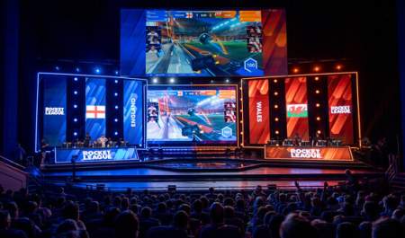 A crowd sit in darkness in a large venue in front of a stage lit in bright blue and red, ready to host an esports event