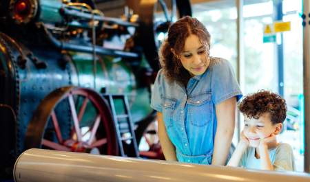 A brother and sister look at a display together at the ThinkTank museum with a steam engine in the background