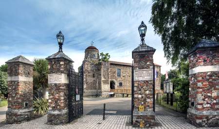 Colchester Castle photographed from outside the Castle Park gates. The Gates are open and four chunky stone pillars with lamps on top sit in the foreground.