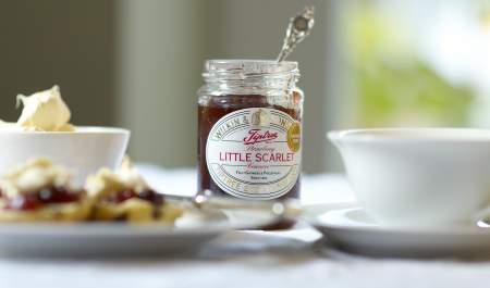 An open jar of Tiptree Littel Scarlet Jam sits on a table beside a bowl of cream, a cup of tea and some scones.
