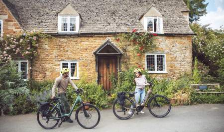 A couple push their bikes past a beautiful old cottage in a Cotswold village