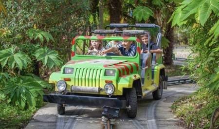 Children ride in a colourful, open-top jungle-themed vehicle on a guided track, surrounded by lush greenery, conveying excitement and adventure.