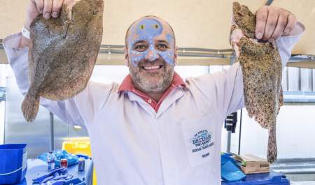Smiling man with face paint holds a large fish in each hand, wearing a white coat with a market logo. Bright, cheerful market setting.