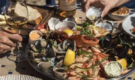 A seafood platter with clams, mussels, prawns, and lemon wedges on a sunny table. Hands holding a wine glass and squeezing lemon over an oyster.