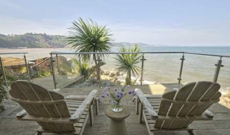 Two wooden lounge chairs on a deck overlook a serene sea view, framed by tropical plants and a glass railing, with a vase of purple flowers on a small table between them.