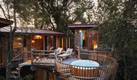 Treehouse retreat at sunset with large windows, cosy interior lighting, chaise lounges, and a hot tub on a wooden deck surrounded by lush trees.