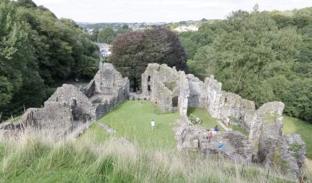 Ancient stone ruins surrounded by lush green trees and grass, with people exploring the site. The scene conveys a sense of history and tranquillity.
