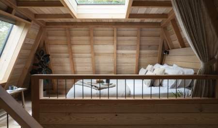 A cosy attic bedroom with wooden walls and ceiling, featuring a skylight above a neatly made bed with cushions. Soft natural light creates a warm ambiance.