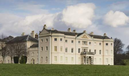 A grand historic Saltram House, cream-colored walls and tall windows set against a blue sky with fluffy clouds, surrounded by green lawns and leafless trees.