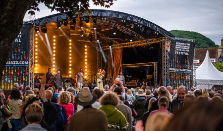 Outdoor music festival scene with a lively crowd watching performers on a lit stage. Banners read "Sidmouth Jazz and Blues Festival." Trees frame the view.