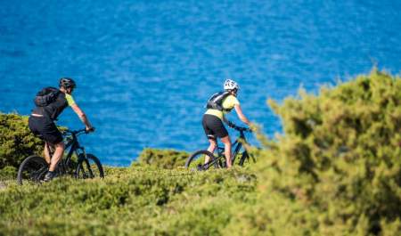 Two cyclists in helmets ride mountain bikes on a grassy trail beside a vivid blue sea, with green shrubs in the foreground, evoking an adventurous spirit.