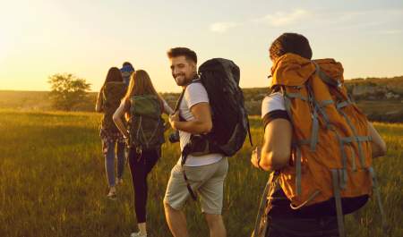 Four hikers with backpacks walk through a field at sunset. One glances back smiling, conveying a sense of adventure.