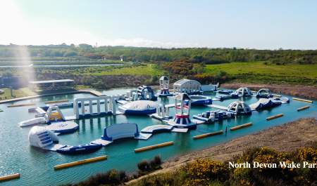 Inflatable water obstacle course on a calm lake, surrounded by lush greenery and bright sunlight. The scene feels adventurous and lively.
