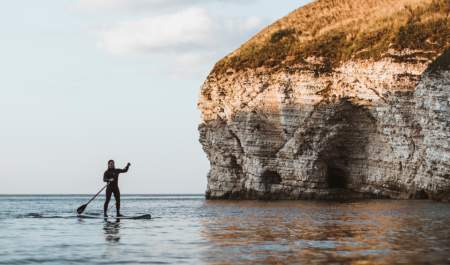 A man paddle boarding in the sea, with the sun appearing on the cliff edge at North Landing, Flamborough