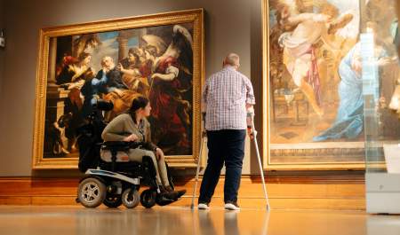 A woman in a wheelchair and a man using crutches viewing two large paintings inside Ferens Art Gallery. The spacious gallery features well-lit artwork and an accessible environment.