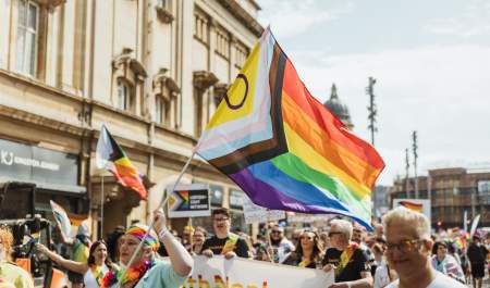 Pride In Hull's parade walking down Carr Lane. The sun is shining, the crowd is smiling and dressed in shorts and t-shirts. A group of people holding a rainbow Smith+Nephew banner are in the foreground with a brightly dressed woman waving an Intersex Inclusive pride flag. Many other pride flags can be seen in the background.