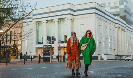 Hull New Theatre in the winter, the white and blue building is light by vertical lights up the colums. Two ladies are walking away after seeing a show. They are bundled up in hats and coats. The lady on the left has blonde hair and is wearing a rusy red fur coat and a beige hat. The lady on the right is wearing a green coat and has red hai and glasses.