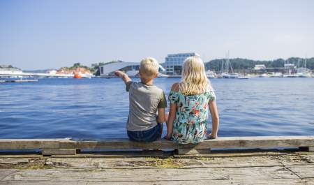 Two children sitting on a dock, pointing towards a building