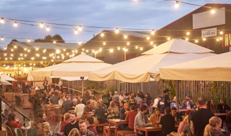 Outdoor dining area with long tables, string lights above, and large umbrellas. People are enjoying food and drinks at dusk, creating a lively and warm atmosphere.
