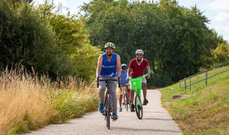People on bikes cycling along a path at Walthamstow Wetlands. Cyclists with protective cycling helmets on. Grass  and foliage along the path. Large trees and greenery in the background.