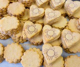 Piles of biscochito cookies, some with a traditional rounded shape and some in a heart shape branded with a Zia symbol.