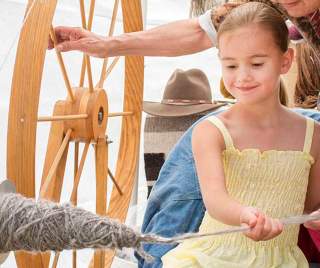 A young person holds yarn coming from a loom with a larger person standing behind them and guiding their hand