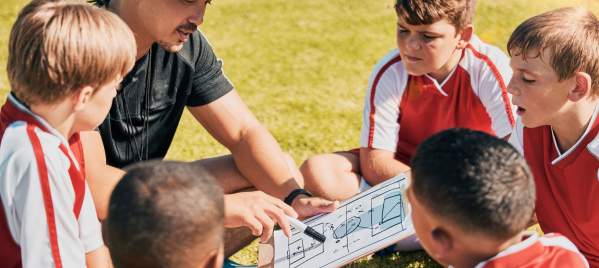 Coach sitting on field with playbook surrounded by youth players.