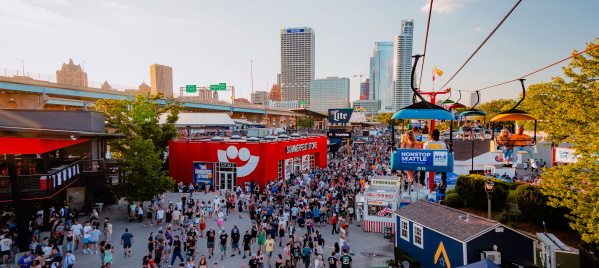 A large crowd moves through the bustling Summerfest grounds in Milwaukee during the day, with the downtown skyline visible in the background. A red building with the Summerfest smiley-face logo serves as the festival store. Above, colorful gondolas on the Skyglider transport people across the venue. The scene is filled with booths, food stands, and signage under a clear sky.