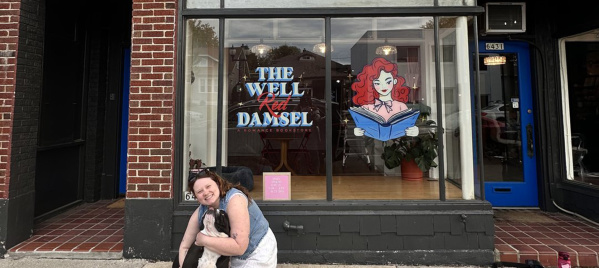 A woman smiles while hugging a black and white dog outside The Well Red Damsel, a romance bookstore with a large window sign featuring a cartoon red-haired woman reading a book.