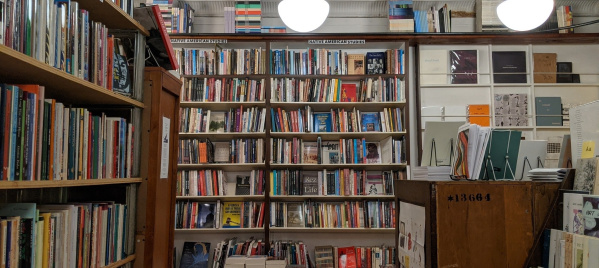 Interior of a bookstore with shelves filled with books, including a section labeled “Native American Studies,” under hanging globe lights, and a wooden display table with books and journals in the foreground.